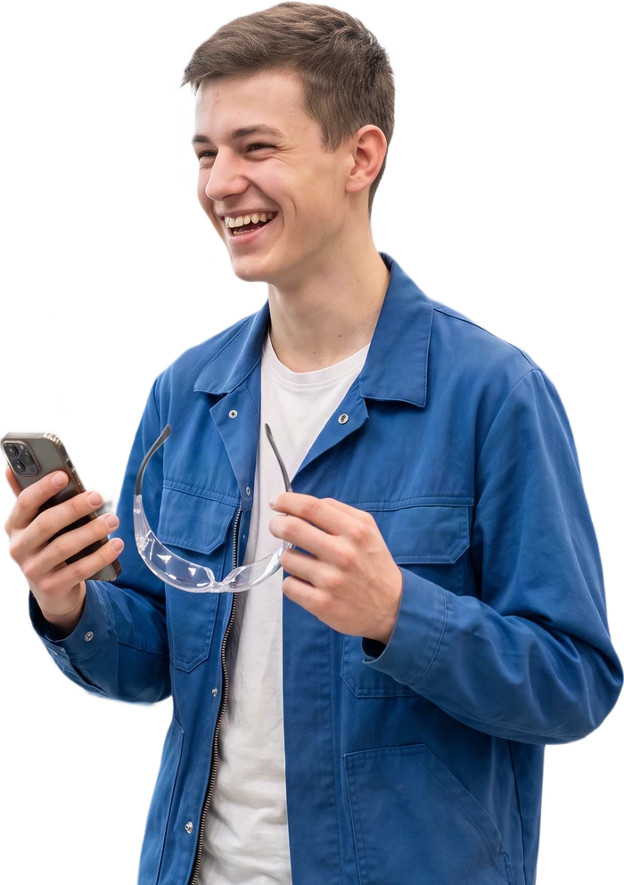 Auszubildender in der Industrie lacht zur Seite, das Handy in der einen, die Schutzbrille in der anderen Hand